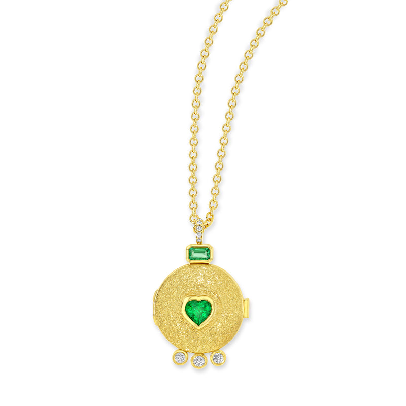 18k yellow gold locket necklace with pavé set white diamond bail. Attaching the bail to the locket is a baguette cut emerald. There is a heart shaped emerald in the center of the locket. At the bottom of the locket are 3 bezel set white diamonds.  