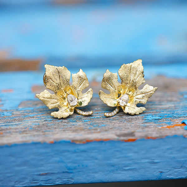 18k yellow gold maple leaf shape stud earings with pear shaped set diamond and pavé diamonds on the leaf stem sitting on a blue wood table